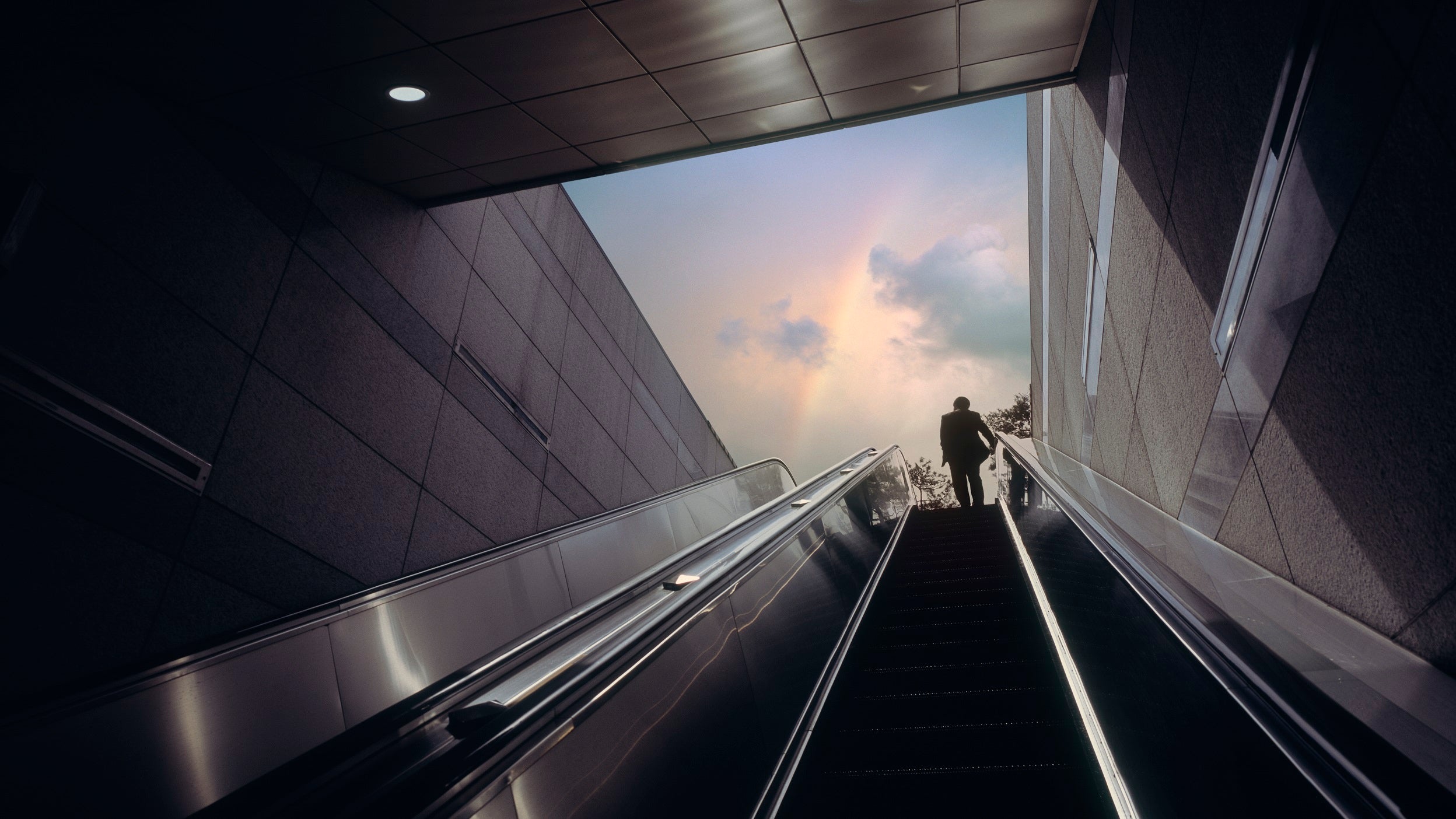 View from behind looking up an escalator as a person emerges from underground with a rainbow viewable in the sky.