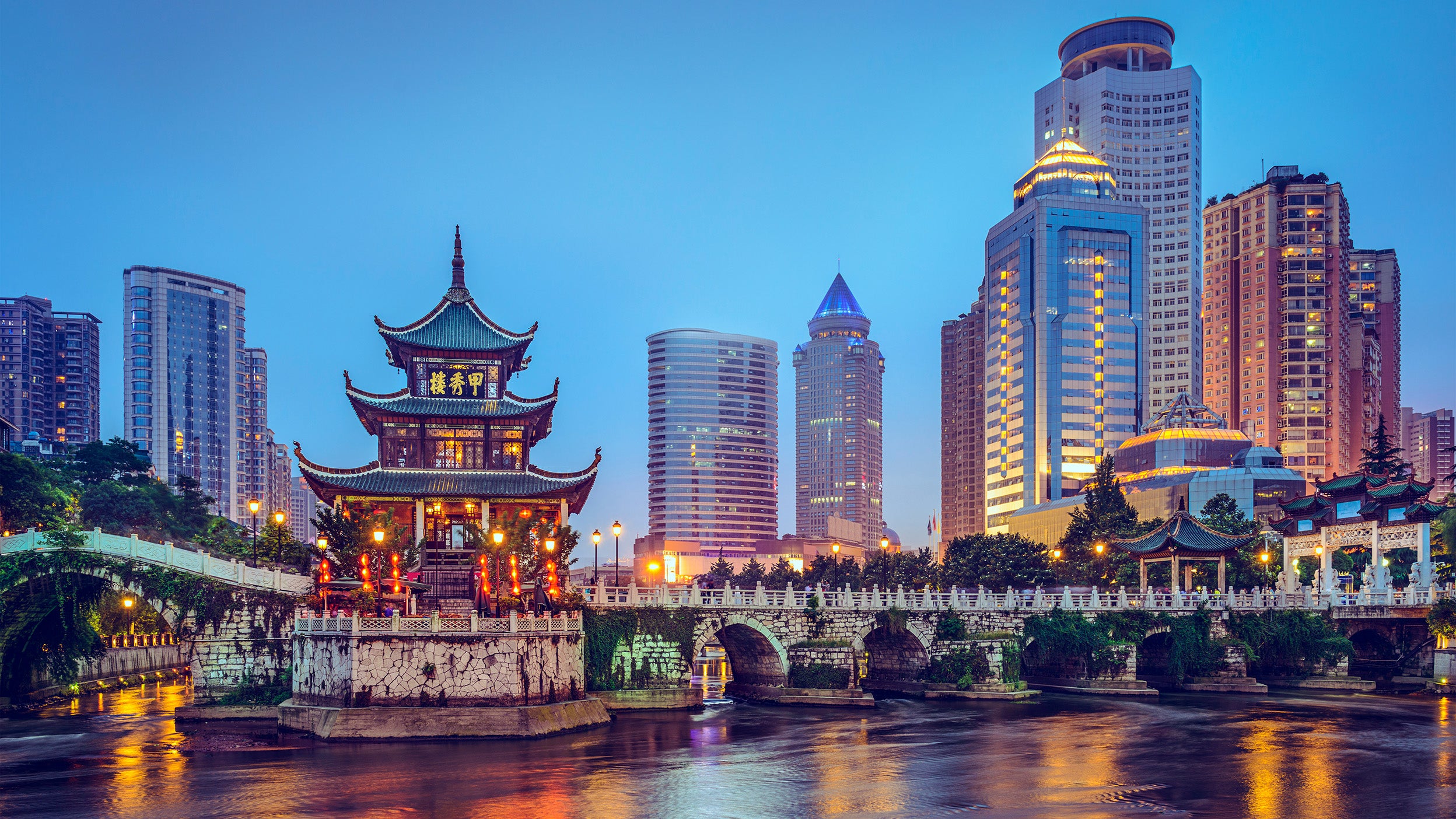 Evening cityscape of Guiyang, China, highlighting the illuminated Jiaxiu Pavilion on a small island connected by an ancient stone bridge, with modern skyscrapers in the twilight background.