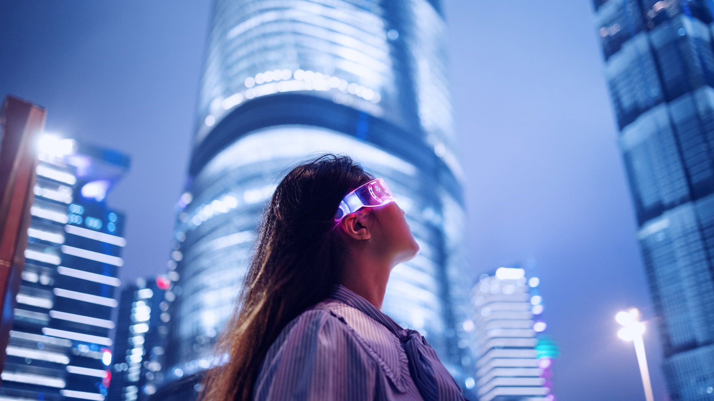Eine Person mit leuchtender Brille blickt in einer nächtlichen Stadtlandschaft zu einem beleuchteten Wolkenkratzer auf.