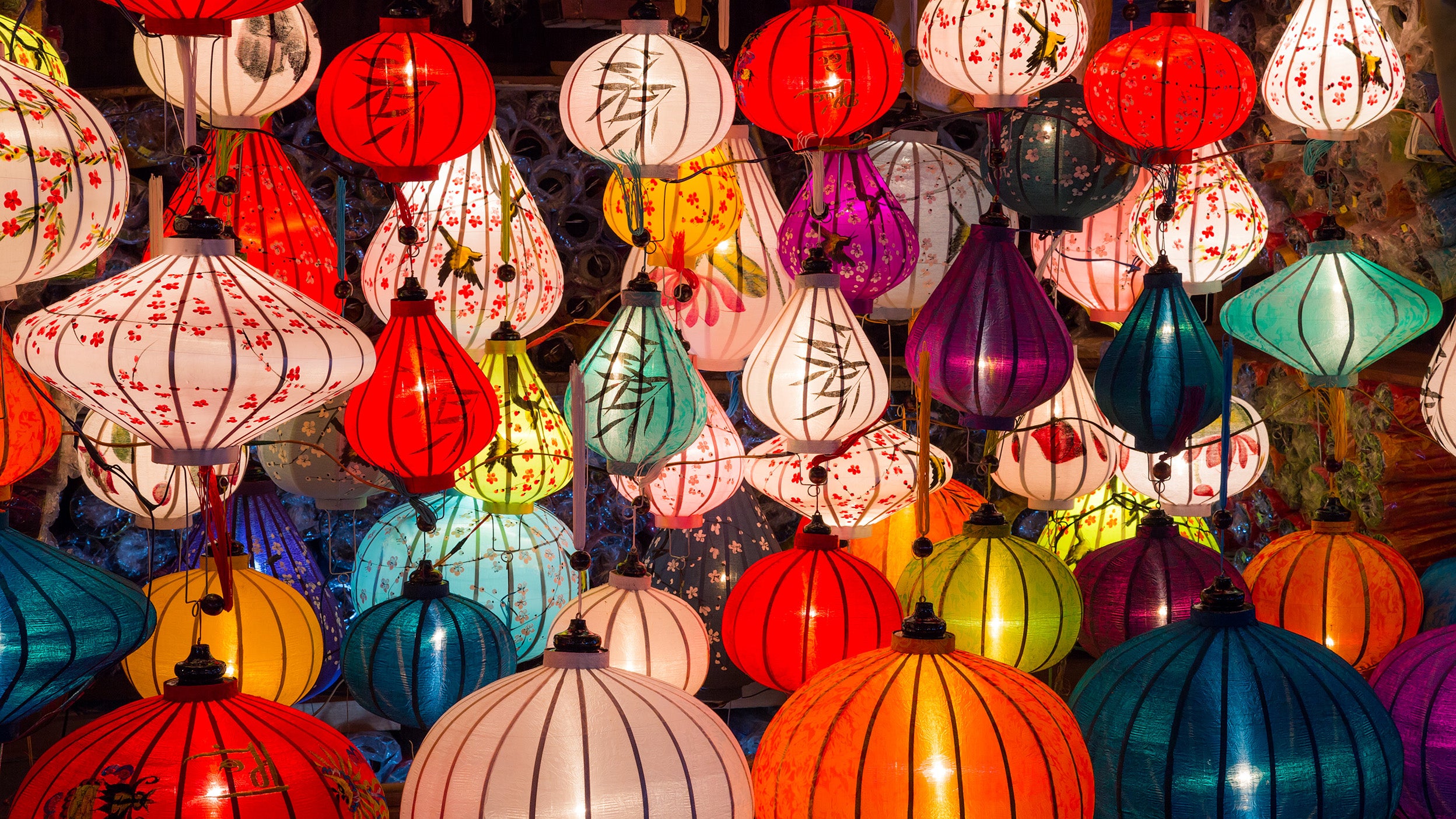 A vibrant display of colourful hanging lanterns in various shapes and sizes, illuminated against a dark background. The lanterns feature intricate designs, including floral patterns and calligraphy, creating a warm and festive atmosphere.