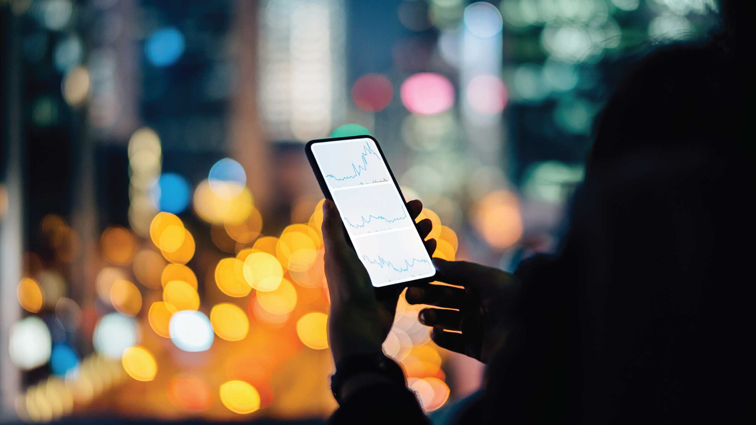 Woman checking financial stock market analysis on smartphone in city, with illuminated city street light and urban skyscrapers as background at night