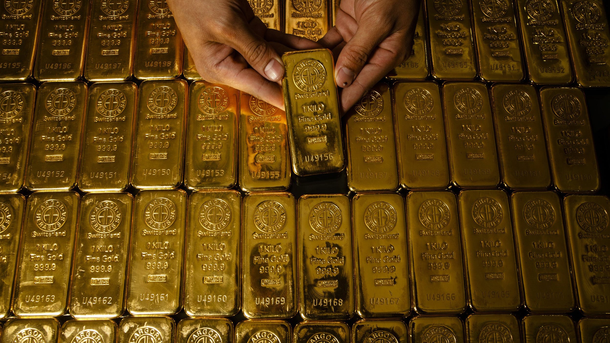 A person holding a gold bar among rows of gold bars, each marked with '1000g' and '999
