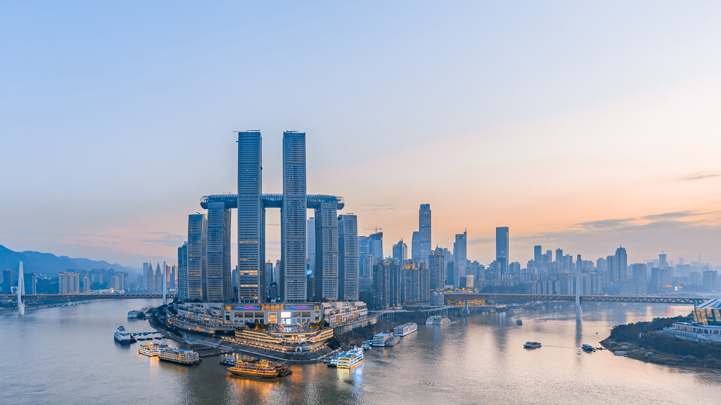 Panoramic view of Chongqing’s skyline at sunset, featuring the Raffles City complex with its connected skyscrapers and skybridge at the confluence of two rivers. Boats navigate the waters, with mountains in the background.