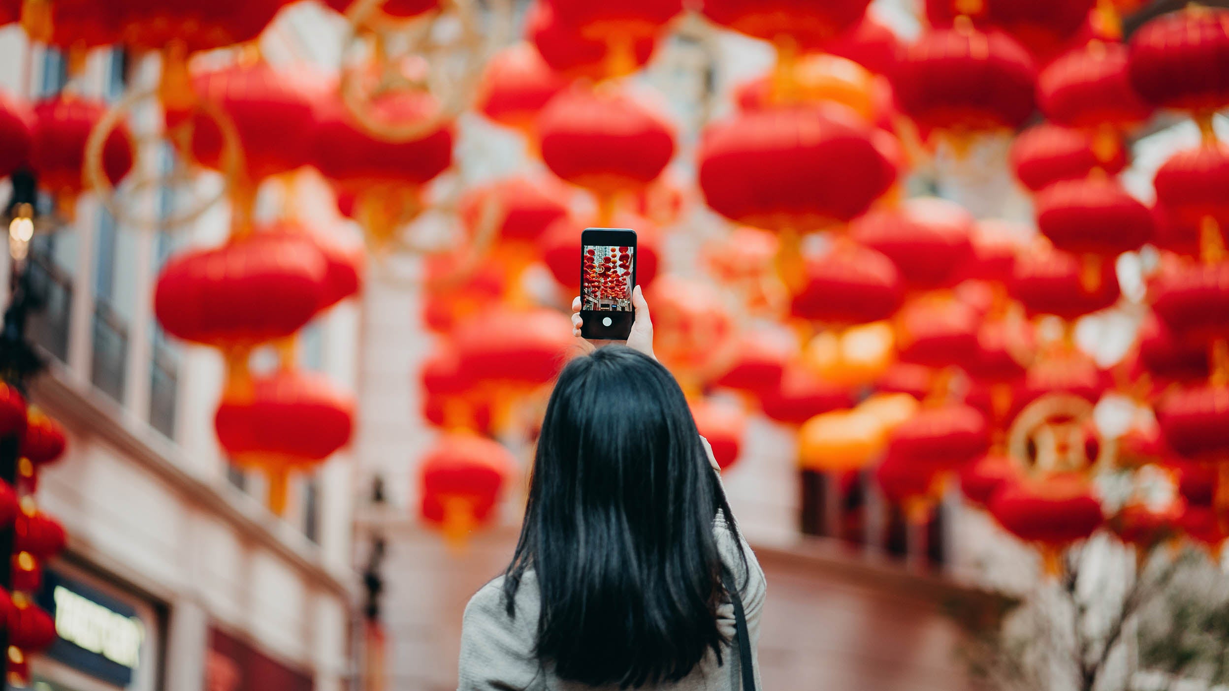 Rear view of woman taking photos of traditional Chinese red lanterns with smartphone on city street.