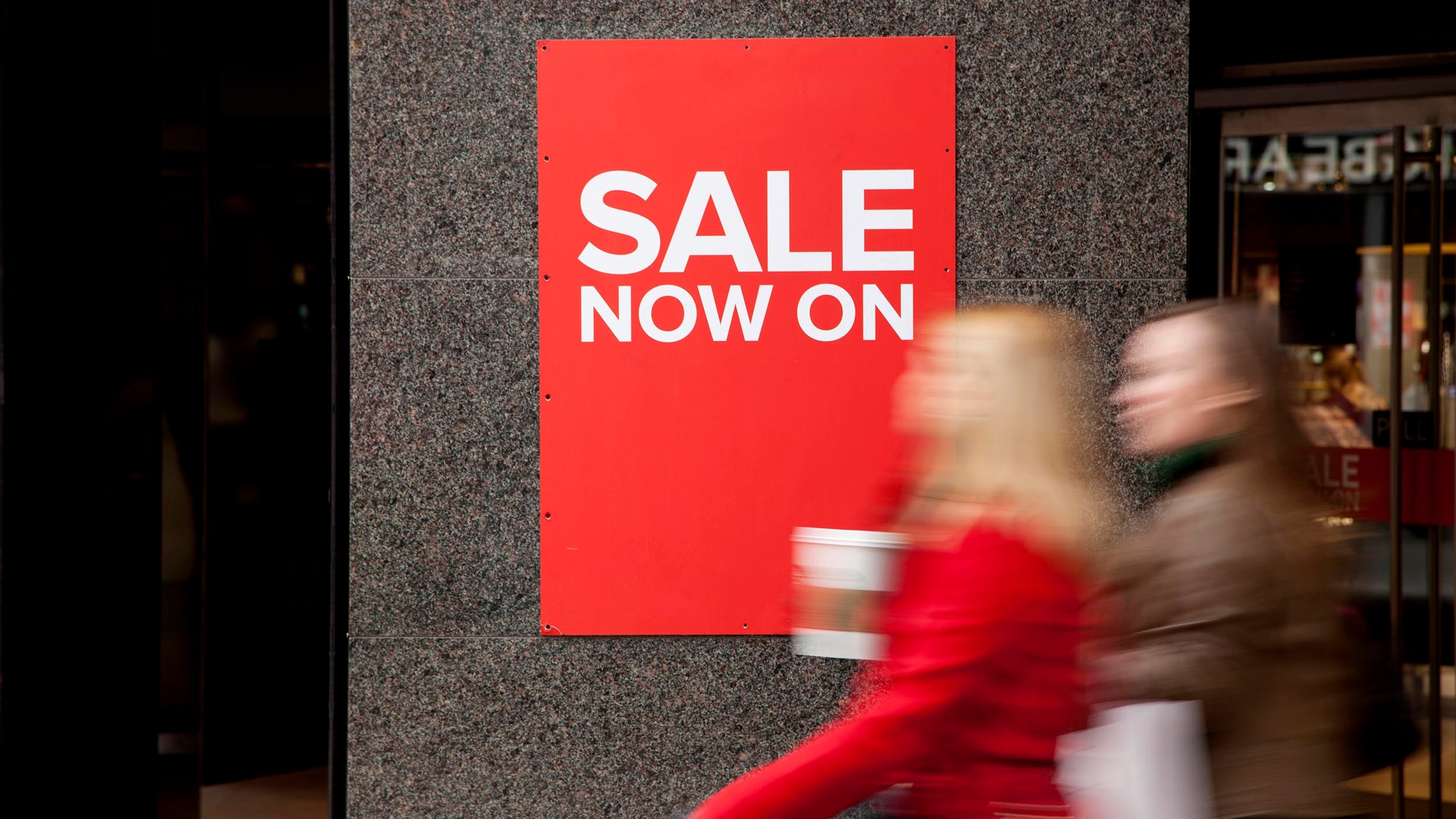 Two people walking past large SALE sign on the exterior of a department store.