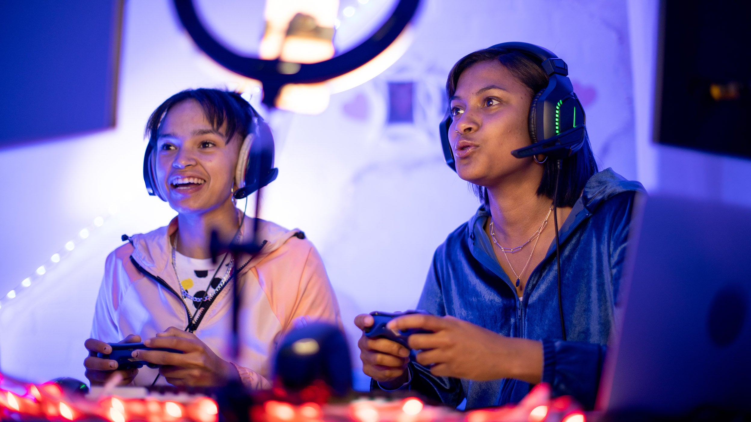 Two girls wearing headphones are holding controllers and focused on playing a video game together.