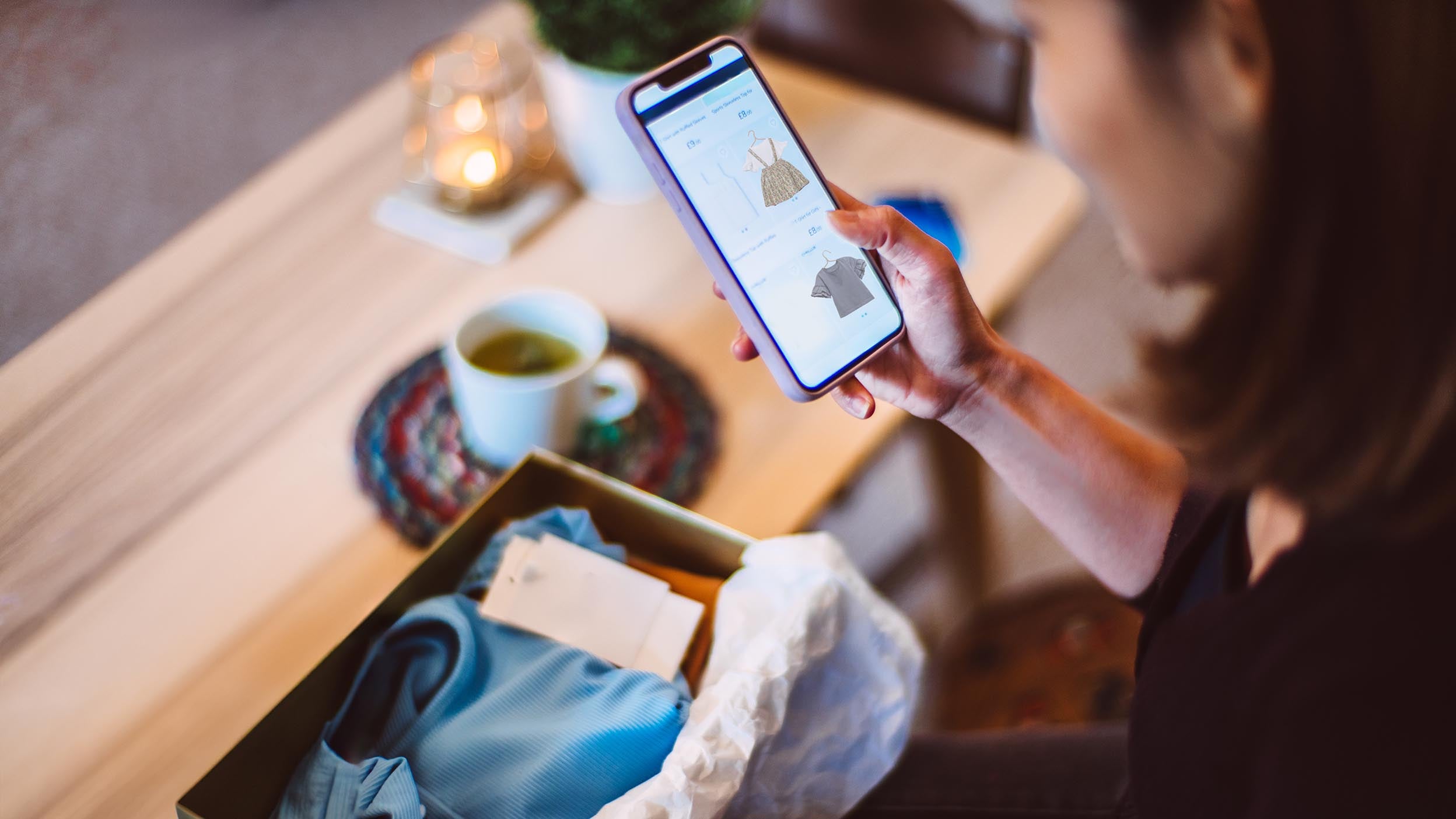 A woman stands over a box of newly arrived clothes, shopping for more items on her smartphone. Invesco QQQ ETF powers e-commerce through many of it’s holding companies. 