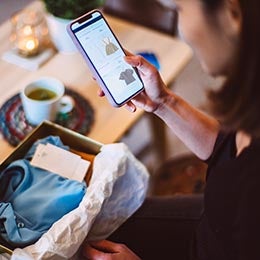 A woman stands over a box of newly arrived clothes, shopping for more items on her smartphone. Invesco QQQ ETF powers e-commerce through many of it’s holding companies. 