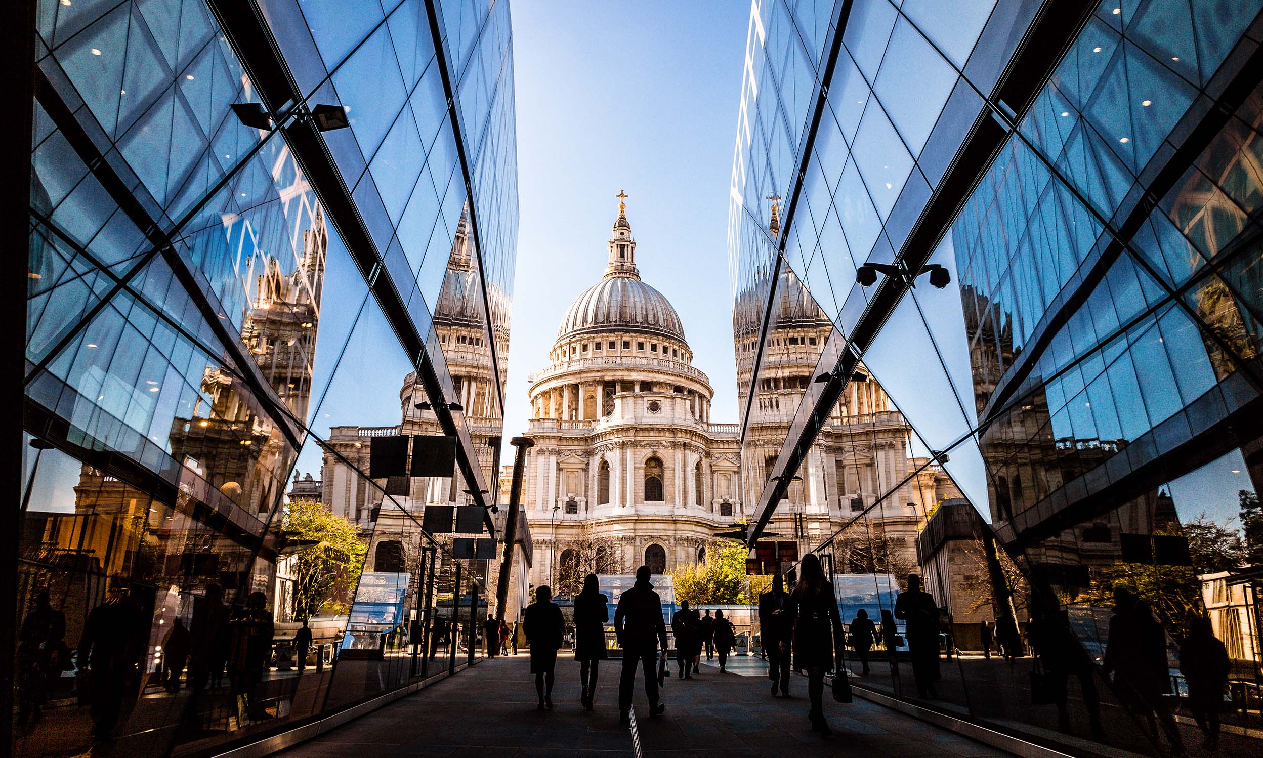 Color image depicting a crowd of people, thrown into silhouette and therefore unrecognizable, walking alongside modern futuristic architecture of glass and steel. In the distance we can see the ancient and iconic dome of St Paul's cathedral.
