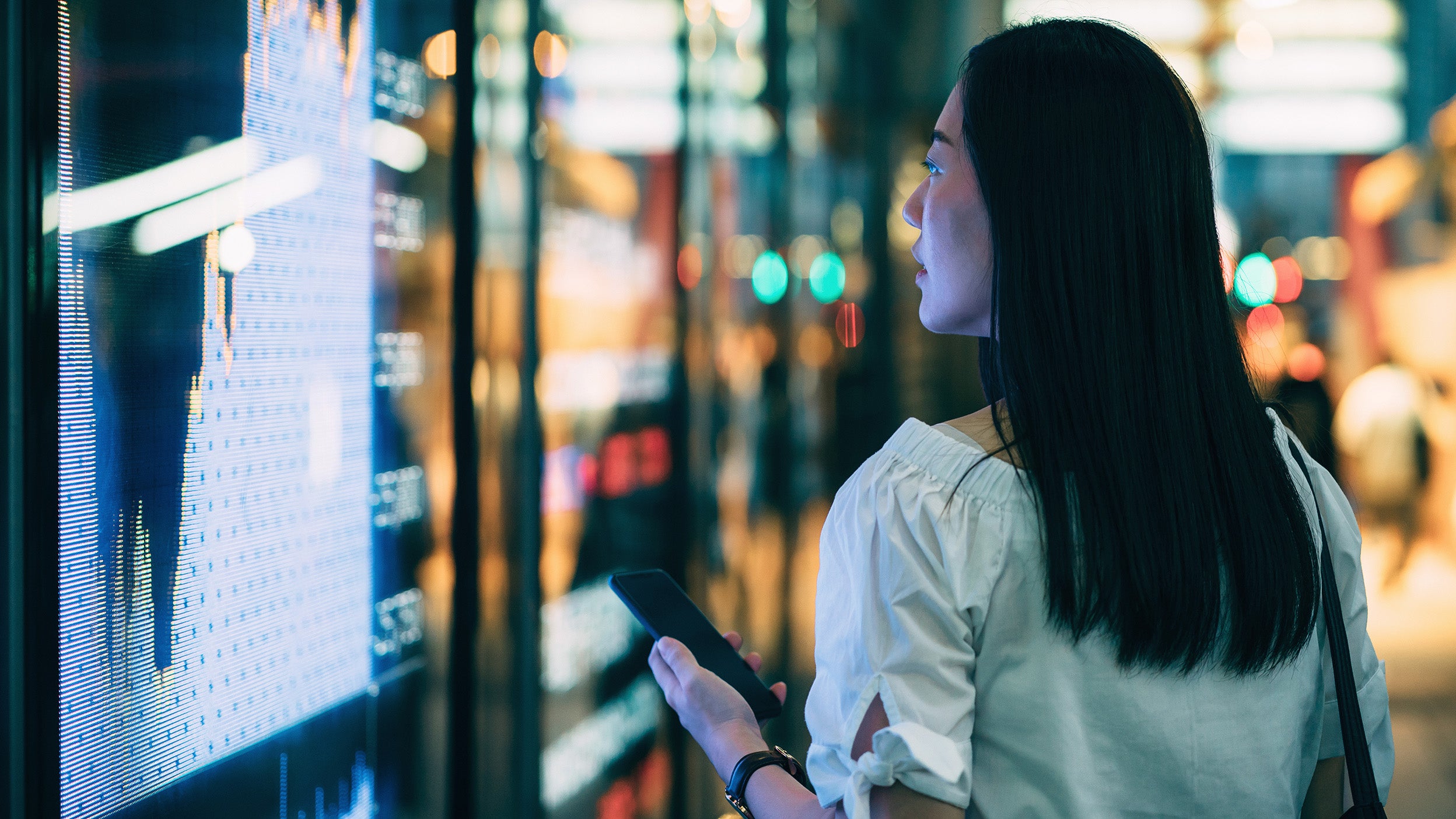 A woman holding a mobile phone looks at a digital screen with market performance.