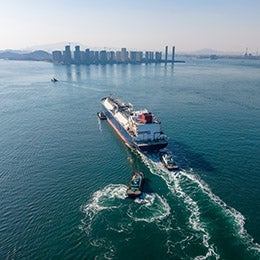 Cargo%20ship%20with%20tugboats%20near%20city%20and%20mountains.